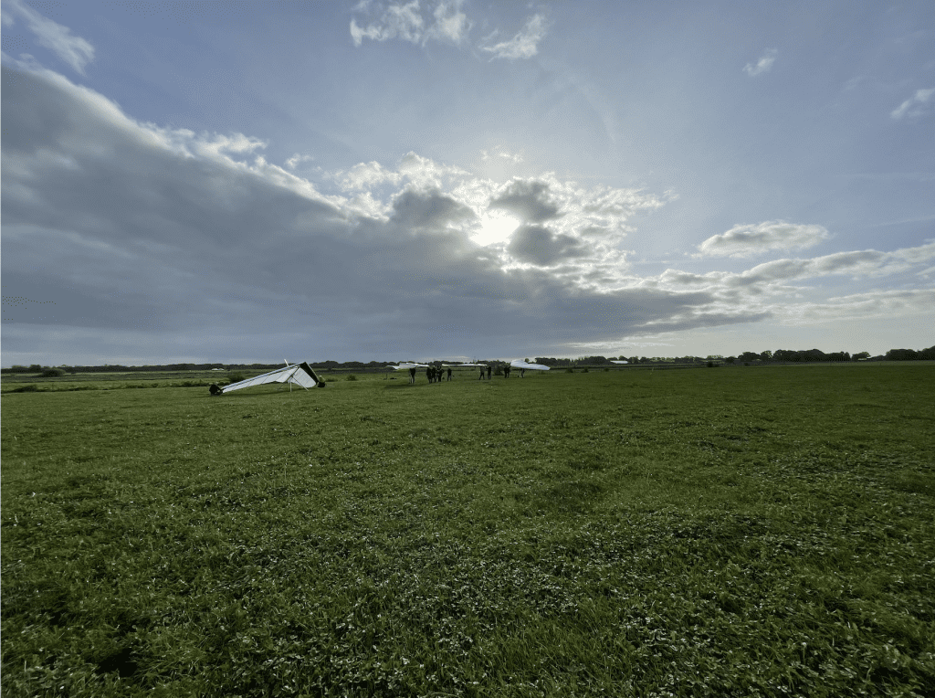 Foto met groothoeklens genomen. Te zien is een breed en leeg weiland met drie deltavleugels en een lucht met wolken waar de zon net doorheen begint te schijnen.